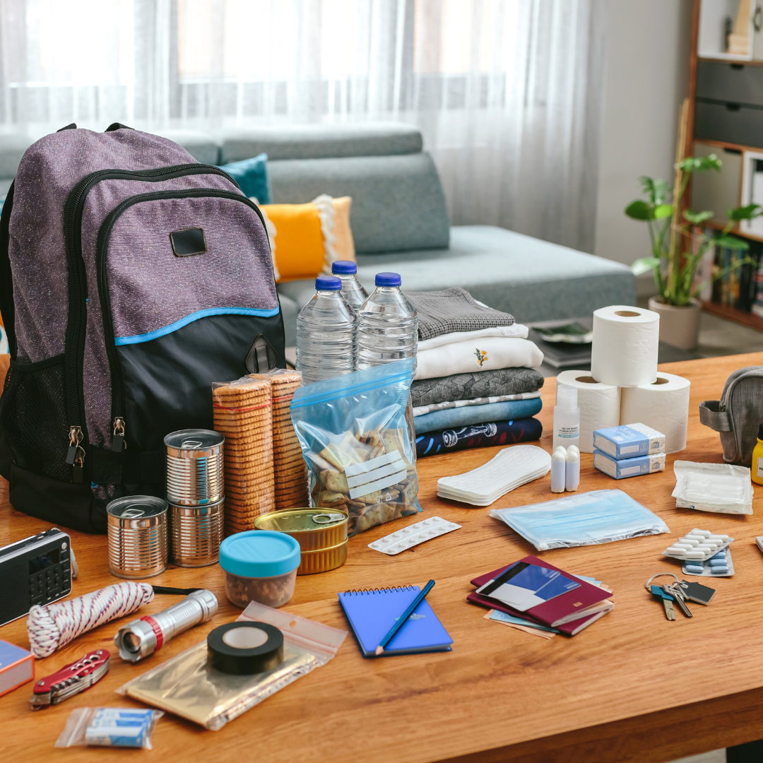 Emergency preparedness backpack with survival gear including bottled water, canned food, rope, flashlight, first aid supplies, face masks, and documents arranged on a table indoors.