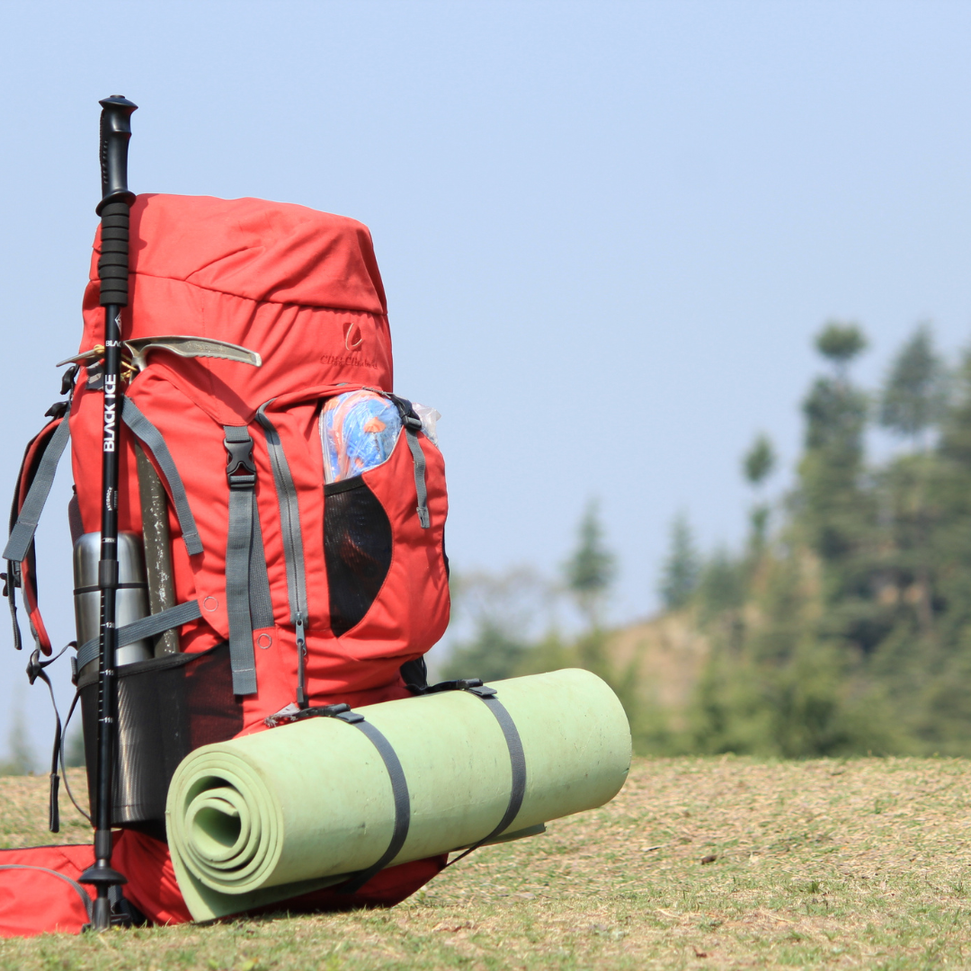 Red survival backpack with hiking pole and rolled sleeping mat placed on grass outdoors for camping or emergency preparedness.