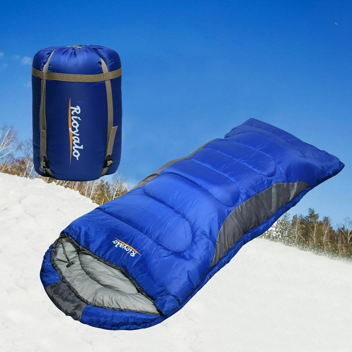 A blue camping sleeping bag and its matching blue carrying bag are shown outdoors on snow under a clear blue sky. The sleeping bag has gray side panels and a gray interior, and the bag has tan straps and a white ROKA logo.