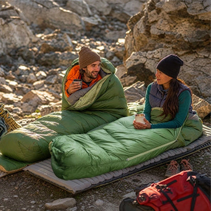 Two people sitting in green camping sleeping bags on an inflatable camping mat in a rocky outdoor mountain setting.