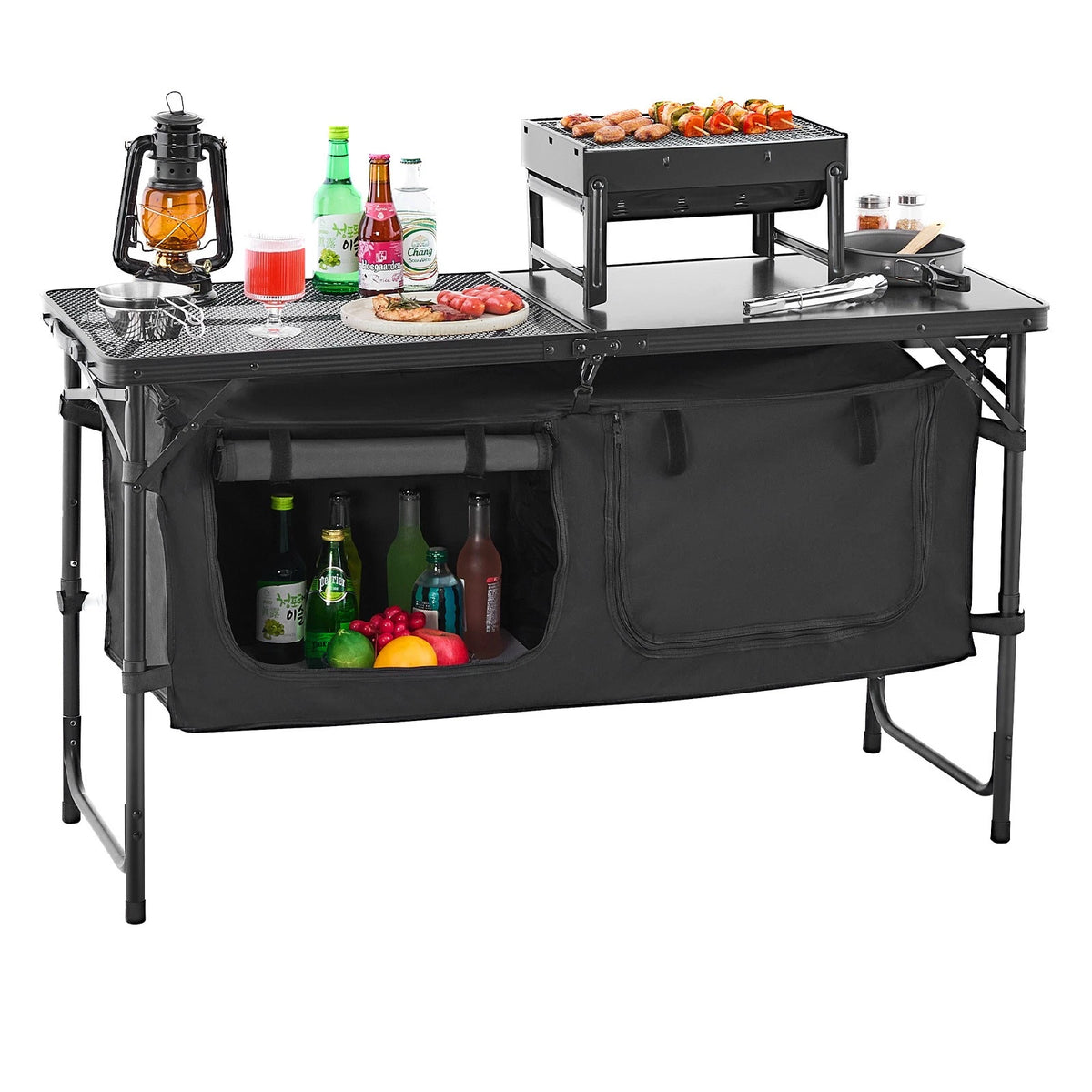 A black folding camp table set up as a cooking station, featuring a tabletop grill with food, condiments, a lantern, and a large front storage compartment filled with bottles and fruit, shown on a white background.