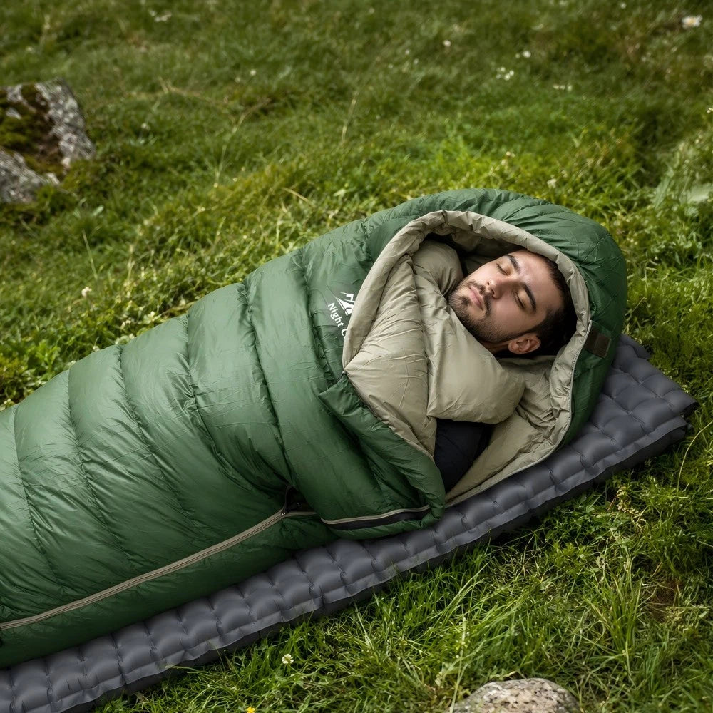 A man sleeping in a green and beige mummy sleeping bag laid out on a dark gray camping mat on a grassy outdoor lawn.